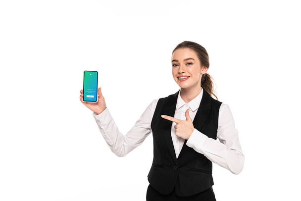 KYIV, UKRAINE - APRIL 7, 2019: happy young waitress pointing with finger at twitter app on smartphone isolated on white - Foto, imagen