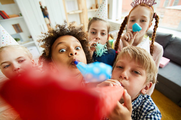 Vista de ángulo alto en grupo multiétnico de niños soplando cuernos de fiesta en la cámara y divirtiéndose
 - Foto, imagen