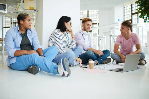 Grupo de jovens colegas inter-raciais pensativos em roupas casuais sentados no chão na biblioteca e trabalhando em projeto criativo juntos
 - Foto, Imagem