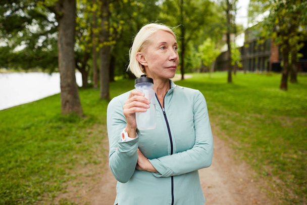 Ξανθιά ώριμη γυναίκα στα ρούχα γυμναστικής έχουν ξεκούραση στο πάρκο και πόσιμο νερό από πλαστικό μπουκάλι, ενώ κινούνται κατά μήκος των δέντρων - Φωτογραφία, εικόνα