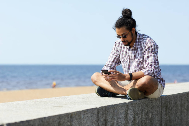 Ritratto integrale dell'uomo latino-americano contemporaneo che si rilassa con lo smartphone seduto sul blocco di cemento via mare, copia lo spazio
 - Foto, immagini