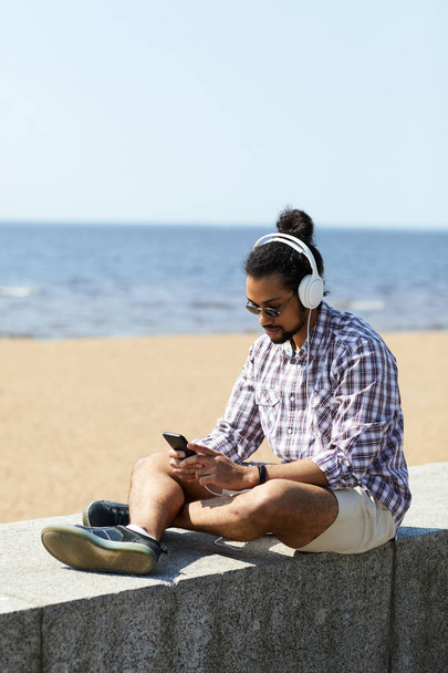 Portrait complet de l'homme latino-américain contemporain se refroidissant sur la plage en écoutant de la musique assis sur un bloc de béton, espace de copie
 - Photo, image