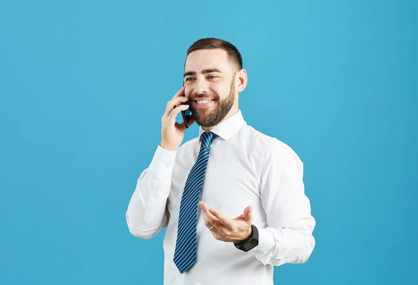 Sonriente joven gerente barbudo optimista en camisa blanca y corbata desnuda de pie contra la pared azul y hablando con el cliente en el teléfono celular
 - Foto, Imagen