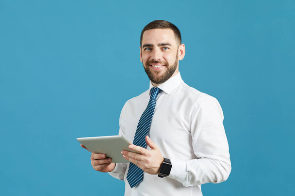 Pozitivní pohledný mladý manažer s bradkou stojící proti modré stěně a používající digitální tablet při pohledu na fotoaparát - Fotografie, Obrázek