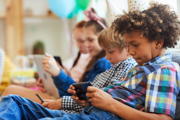 Side view at group of children using smartphones sitting in row at birthday party, copy space - Fotoğraf, Görsel