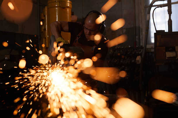 Ouvrier d'usine occupé dans des protecteurs d'oreille debout dans l'atelier et sciant le métal avec la machine de découpe
 - Photo, image