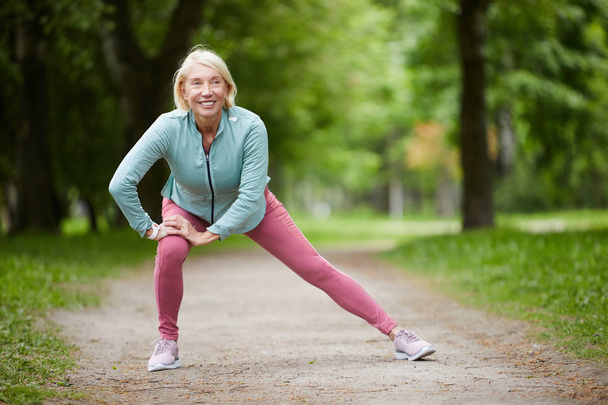 Contemporary mature blonde female in activewear doing exercise for leg stretch while enjoying workout in natural environment - 写真・画像