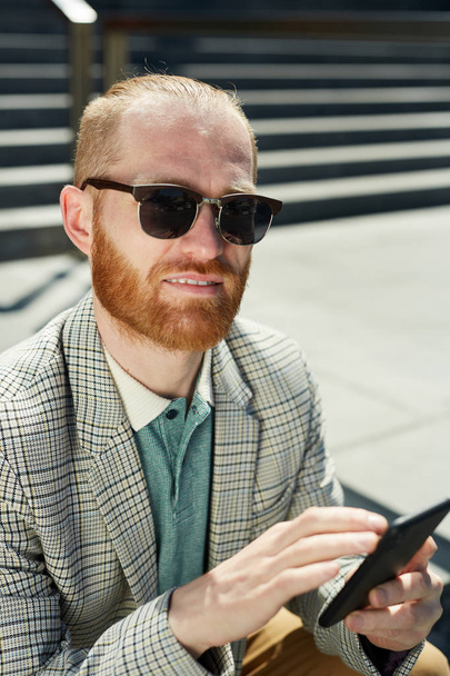 Портрет улыбающегося уверенного хипстера с рыжей бородой в клетчатой куртке, использующего телефон для работы
 - Фото, изображение