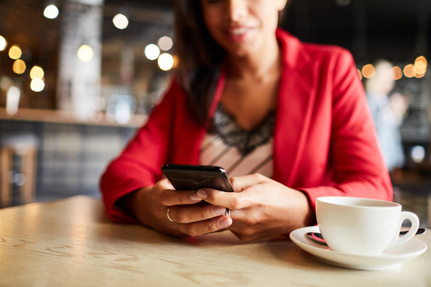 Close-up of positive woman sitting at table with coffee cup in cafe and using smartphone for online communication - Photo, Image