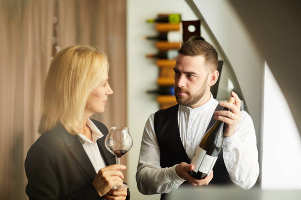 Warm toned portrait of sommelier showing wine bottle to woman in restaurant, copy space - Photo, Image
