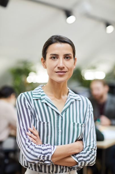Талия портрет успешной бизнес-женщины, смотрящей в камеру, позируя в офисе стоя со скрещенными руками
 - Фото, изображение
