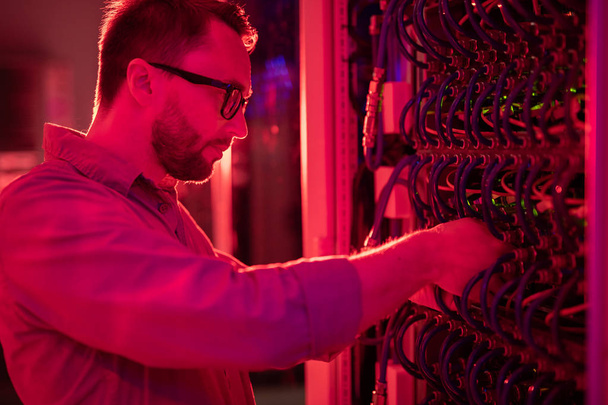 Serieux technicien de réseau barbu concentré dans des lunettes debout à l'armoire de l'ordinateur central et de fixation superordinateur, lumière rouge
 - Photo, image
