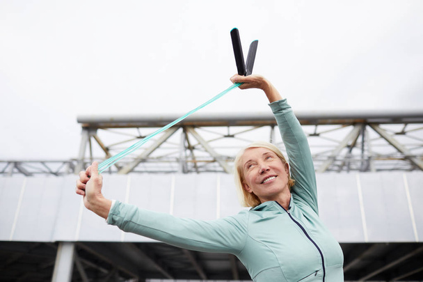 Счастливая взрослая спортсменка в спортивной одежде делает упражнения на гибку с скакалкой, наслаждаясь тренировками в городской среде
 - Фото, изображение