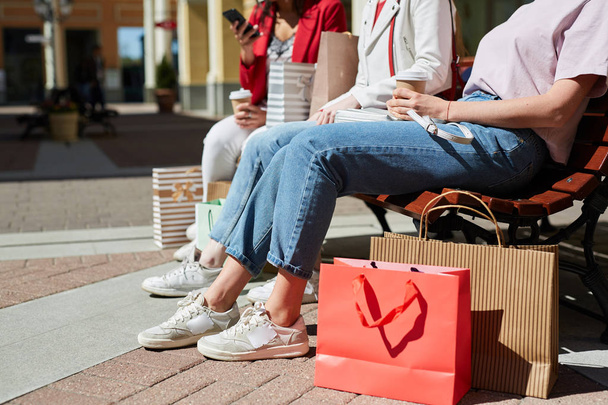 Zbliżenie nierozpoznawalnych kupujących dziewczyna w casual stroje siedzi na ławce na zewnątrz i picie kawy podczas odpoczynku po zakupach - Zdjęcie, obraz