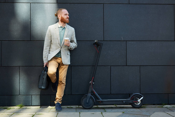 positiv aktiver Hipster junger Geschäftsmann mit Bart lehnt an Hauswand auf der Straße und trinkt Kaffee, moderner Tretroller zum Pendeln - Foto, Bild