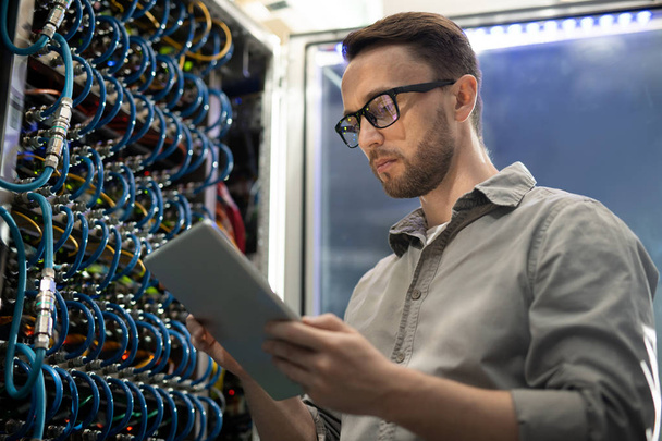 Serious smart young system engineer with beard standing in server room and using tablet while testing connectivity through app - 写真・画像