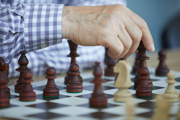 Tanned wrinkled hand of old man in checked purple and white shirt making chess move with dark figures - Foto, afbeelding