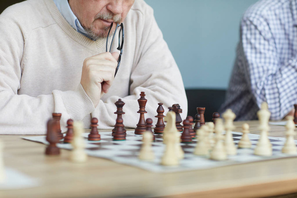 Aged focused experienced chess player with grey beard biting glasses while analyzing chess game - Photo, Image