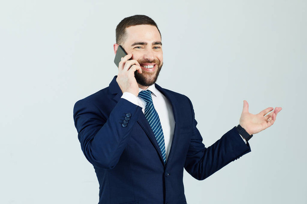 Positive confident young male manager in dark blue suit standing against isolated background and gesturing hand while communicating on mobile phone - Фото, зображення