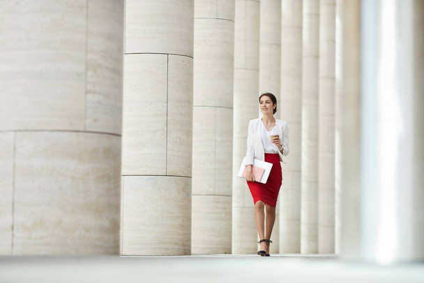 Ganzkörperporträt der schönen Geschäftsfrau mit rotem Rock, die entlang der Säulenreihe auf die Kamera zugeht, Kopierraum - Foto, Bild