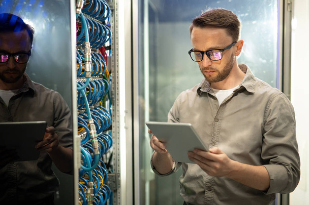 Serious skilled young data center engineer in glasses standing at mainframe and examining cables while doing checkup - 写真・画像