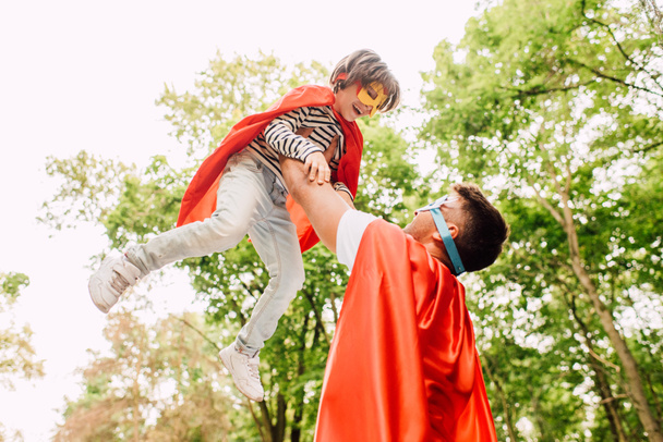 Отец в костюме супергероя держит сына в руках, пока ребенок смеется и смотрит на отца
 - Фото, изображение