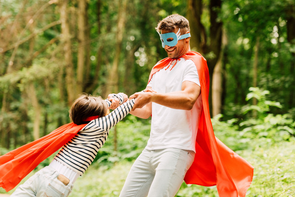 Vater in Mantel und Maske spinnt kleinen Jungen in rotem Superheldenmantel - Foto, Bild