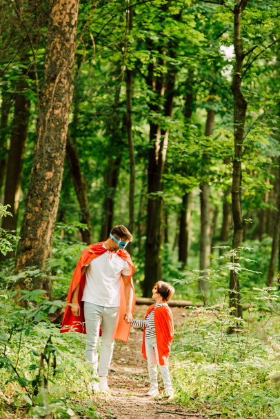 full length view of father and son standing in superhero costumes in forest and looking at each other - Photo, Image
