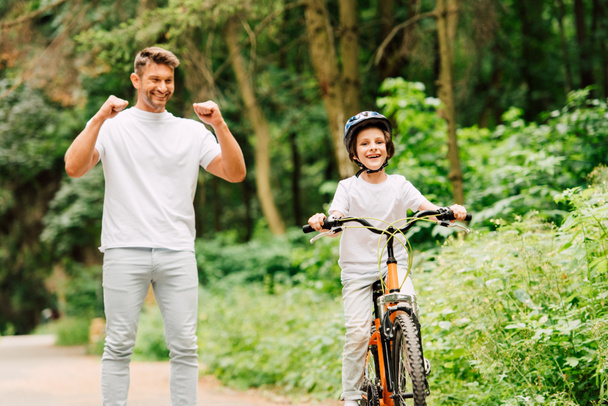 father smiling and cheering son while boy riding bicycle and looking at camera - Foto, Imagem