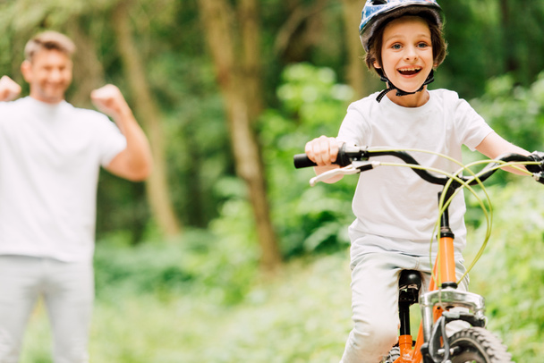 valikoiva painopiste isä hurraavat poika kun poika ratsastus polkupyörän ja katsot kameraa
 - Valokuva, kuva