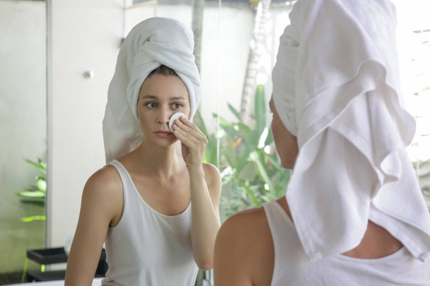 Portrait of a beautiful young woman with a towel on the head standing in the bathroom and cleaning her face with cotton pads in front of the mirror, removing makeup, beauty and spa concept - Photo, Image
