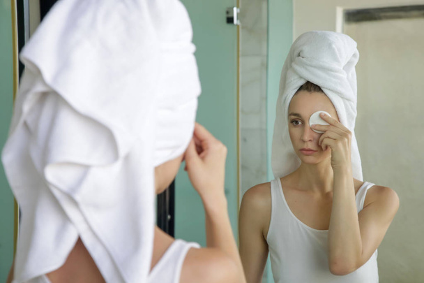 Portrait of a beautiful young woman with a towel on the head standing in the bathroom and cleaning her face with cotton pads in front of the mirror, removing makeup, beauty and spa concept - Photo, image