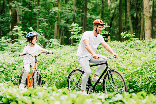 selective focus of father and son smiling while riding bicycles in forest - Φωτογραφία, εικόνα