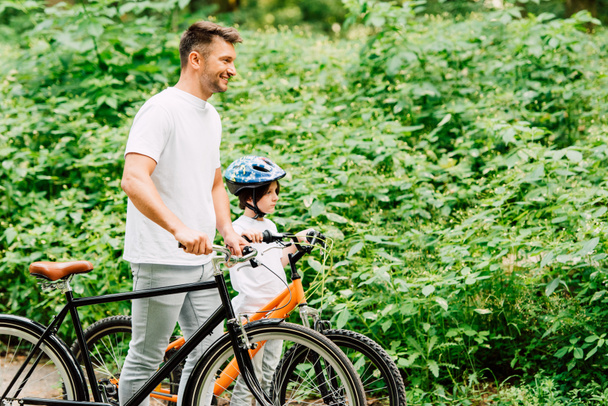 side view of son and father standing with bicycles near forest and looking forward - Foto, Imagem