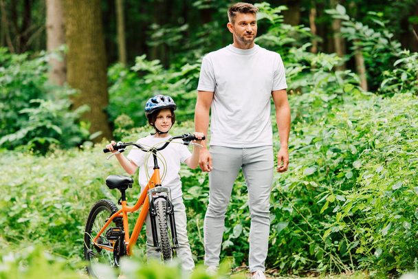 full length view of father and son standing on road near forest and looking forward while son holding bicycle - Foto, immagini