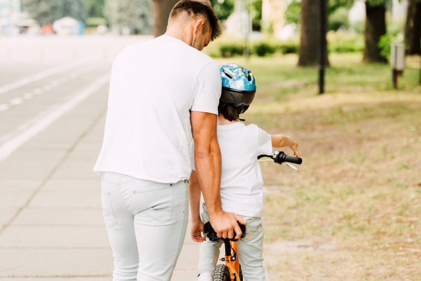 vista trasera del padre ayudando a su hijo a montar en bicicleta mientras niño sentado en bicicleta
 - Foto, Imagen