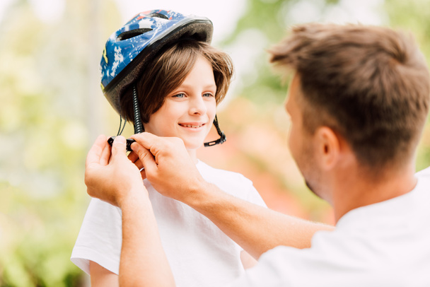 selective focus of happy son looking at dad while father putting helmet - Photo, Image
