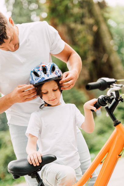 father putting helmet on son while boy trying to sit on bicycle - Foto, afbeelding