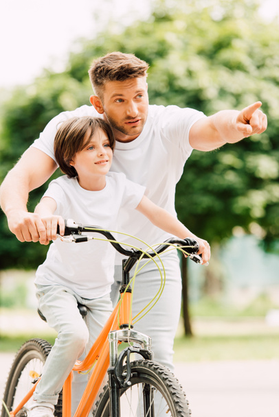 father and son looking forward while kid riding bicycle and dad pointing with finger - Foto, afbeelding