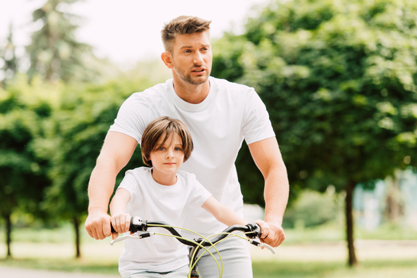 father looking forward while son sitting on bicycle and looking at camera - Φωτογραφία, εικόνα