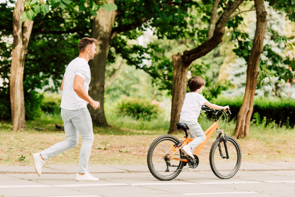 Ganzkörperansicht des Vaters, der dem Sohn hinterherläuft, während das Kind Fahrrad fährt - Foto, Bild