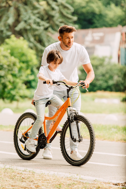 pohled na otce, který se dívá dopředu a pomáhá synovi jezdit na kole, zatímco syn sedí na kole - Fotografie, Obrázek