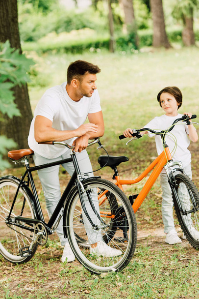 πλήρους μήκους θέα του πατέρα και του γιου που γέρνει στα ποδήλατα, ενώ στέκεται στο πάρκο - Φωτογραφία, εικόνα