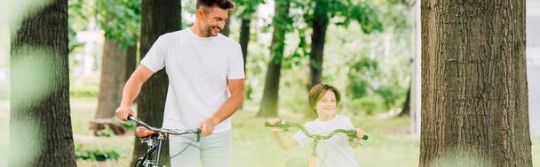 panoramiczny strzał ojca i syna chodzenie w lesie z rowerami i uśmiechnięty podczas tata patrząc na dziecko  - Zdjęcie, obraz