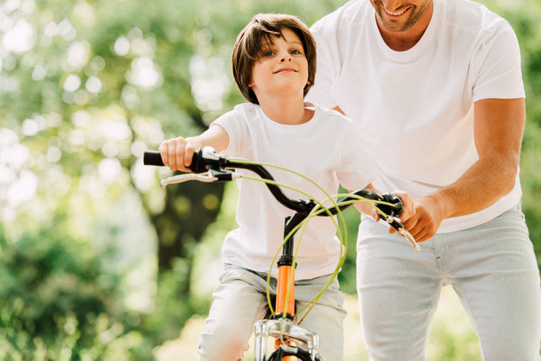 cropped view of father helping son while kid riding bicycle and looking forward  - Фото, изображение