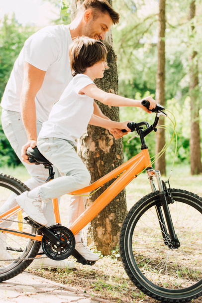 side view of son riding bicycle and father walking next to kid and holding sit of bike  - Fotó, kép