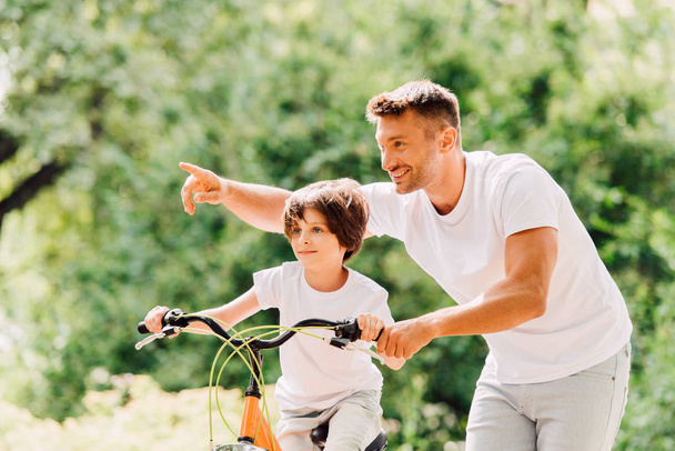 son riding bicycle while father helping kid and pointing with finger  - Foto, Imagen