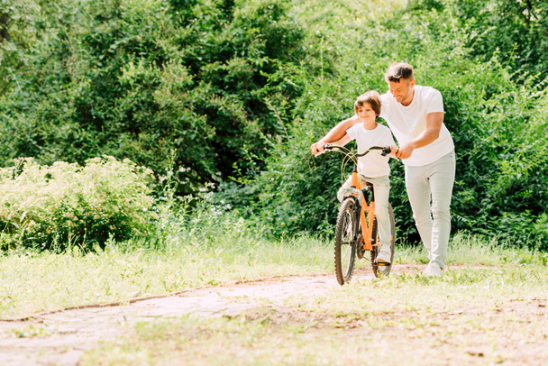 Ganzkörperansicht von Vater hilft Sohn, indem er Fahrradgriffe hält, während Sohn Fahrrad fährt - Foto, Bild