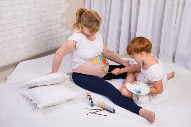 Little red-haired girl draws on the belly of a pregnant mom drawing paints - 写真・画像