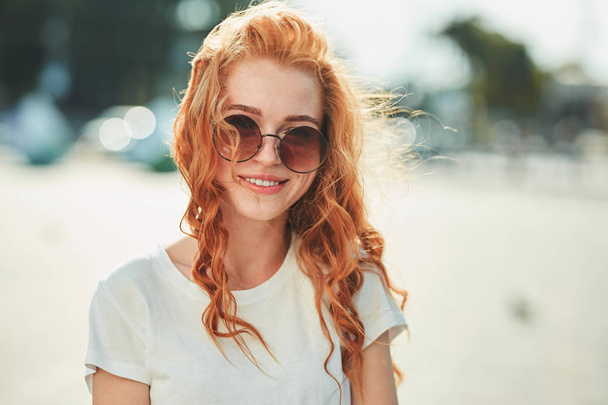 Kaunis punatukkainen tyttö valkoisessa t-paidassa ja aurinkolaseissa kävelee kadulla ja hymyilee kehyksissä.
 - Valokuva, kuva